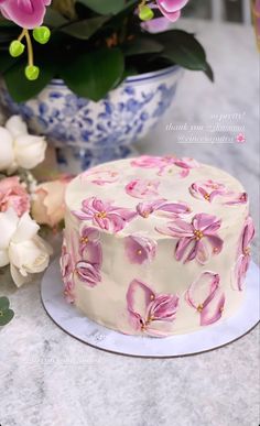 a white cake with pink flowers on it sitting next to a blue and white vase