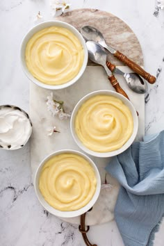 three white bowls filled with yellow pudding on top of a marble counter next to spoons