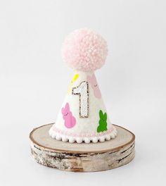 a pink and white birthday hat sitting on top of a piece of wood with pom - poms