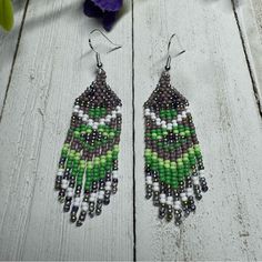 green and white beaded earrings sitting on top of a wooden table next to purple flowers