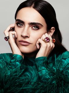 a woman with green feathers and rings on her fingers, posing for a photo in front of a gray background