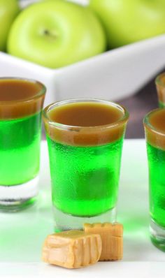 three shot glasses filled with green liquid next to some pieces of candy and apples in the background