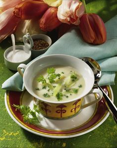 a bowl of soup is sitting on a plate with spoons next to it and flowers in the background