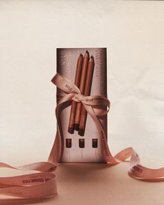 the packaging is wrapped in pink ribbon and has three brown pencils on top of it