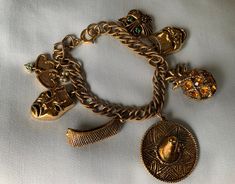 "Stunning gold-toned charm bracelet.  There are 7 charms, all gold toned. They include: Linked hearts Comedy mask Tragedy mask Canoe Sombrero Pineapple (missing one jewel) Cat Length: 7.5\" Very good condition. Normal wear." Gold Charms Jewelry As Fashion Accessory, Adjustable Gold Bracelets With Vintage Charm, Gold Bohemian Jewelry For Souvenir, Adjustable Gold Charm Bracelet, Gold Charm Bracelet As A Souvenir, Vintage Gold Bracelets, Adjustable Gold Bracelet With Vintage Charm, Adjustable Gold Charm Bracelet Collectible, Gold Vintage Bracelets