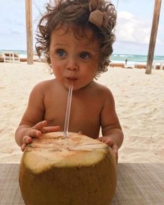 a baby sitting on the beach with a straw in it's mouth and drinking from a coconut