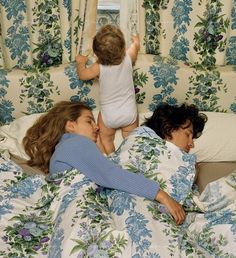 two women and a baby are laying in bed with blue flowers on the walls behind them