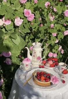 a table topped with plates and cups filled with fruit next to pink flowered bushes