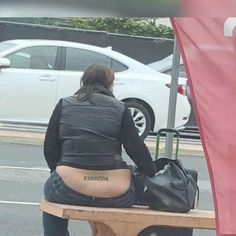 a woman sitting on top of a wooden bench next to a white car and holding a black bag