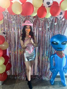 a woman standing in front of balloons with an alien figure next to her holding a lit candle