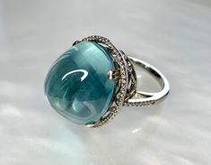 A captivating and luxurious sea- foam green aquamarine piece reminiscent of the first steps we take into the clear blue ocean waters. This super sized cocktail ring featuring a high domed 24.21 carat cabochon center stone surrounded by a halo of diamonds weighing 0.70 carats. Set in 18k white gold, this collector's item boasts rare geometric shaped cut- out detailing on the gallery beneath the stone. *Approximate stone measurements: Height: 19 mm Width: 16 mm Approximate ring weight: 13.92 grams Luxury Oval Cabochon Birthstone Ring, Luxury Blue Crystal Ring With Center Stone, Luxury Blue Crystal Ring In Sterling Silver, Blue Crystal Ring In Fine Jewelry Style, Luxury Oval Cabochon Rings For May Birthstone, Luxury Diamond Gemstone Cabochons, Luxury Oval Cabochon May Birthstone Rings, Luxury Crystal Ring With Stone Setting For Formal Occasions, Luxury Oval Cabochon Rings With Gemstone Accents