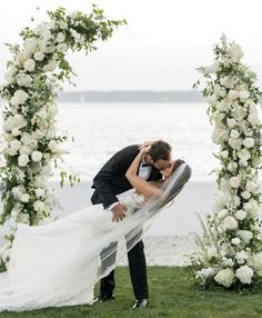 a bride and groom kissing in front of an arch with flowers on the grass by the water