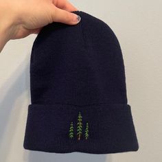 a hand holding onto a blue hat with trees embroidered on the front and back side