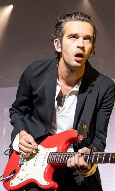 a man holding a red guitar in his right hand and wearing a black suit jacket