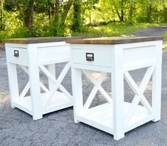 two white side tables sitting next to each other on gravel ground with trees in the background