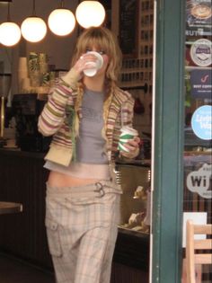 a woman standing in front of a store drinking from a cup while wearing plaid pants