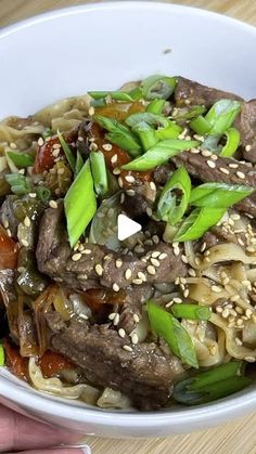 a white bowl filled with noodles and meat on top of a wooden table next to a person's hand