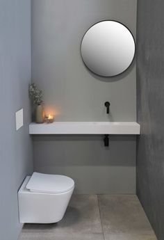 a white toilet sitting under a round mirror next to a sink in a room with grey walls