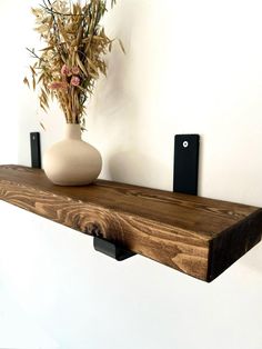 a wooden shelf with two black brackets on it and a vase filled with flowers in the background