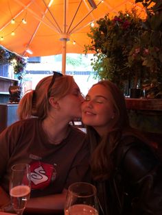 two women sitting at a table kissing each other with an umbrella in the back ground