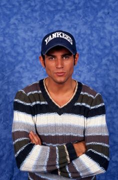 a young man wearing a yankees hat and striped sweater poses for a photo in front of a blue backdrop