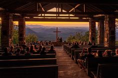 people are sitting in pews and watching the sun set over mountains with a cross on top