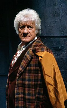 an older man with white hair wearing a brown and yellow coat, standing in front of a black wall