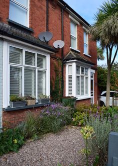 a brick house with white shutters and flowers in the front yard, next to a gravel path