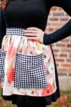 a woman wearing a black and white checkered apron with buttons on the side, standing in front of a brick wall