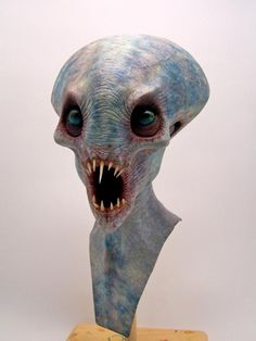 an alien head with blue eyes and mouth is on a wooden stand against a white wall