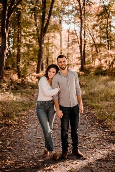 a man and woman holding hands while standing on a path in the woods with trees