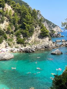 people are swimming in the clear blue water near some rocks and trees on the shore