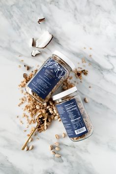 two jars filled with nuts on top of a white marble counter next to a spoon