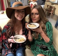 two women sitting on a couch holding plates of food in their hands and smiling at the camera