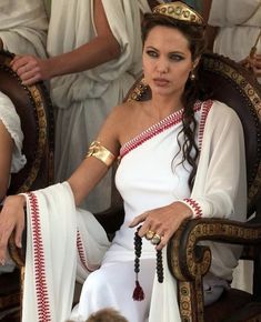 a woman sitting in a chair with other women behind her wearing white dresses and gold jewelry