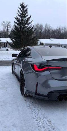 the rear end of a silver sports car parked on snow covered ground in front of a house