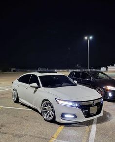 two cars are parked in a parking lot at night, one is white and the other is black