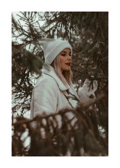 a woman wearing a white coat and hat standing next to a pine tree in the snow