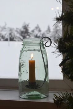 a lit candle in a jar on a window sill next to a pine tree