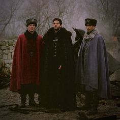 three men standing next to each other in front of a stone wall with trees behind them