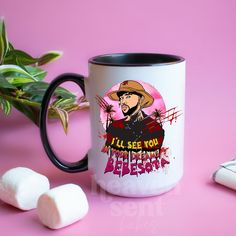 a white and black coffee mug sitting on top of a pink table next to marshmallows