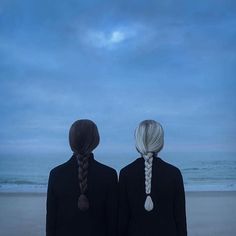 two people standing on the beach looking out at the ocean with their hair in braids