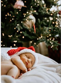 a baby wearing a santa hat sleeping in front of a christmas tree with lights on it