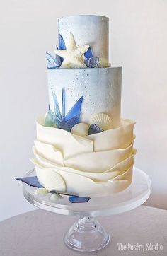 a three tiered cake decorated with blue and white sea shells on a clear plate