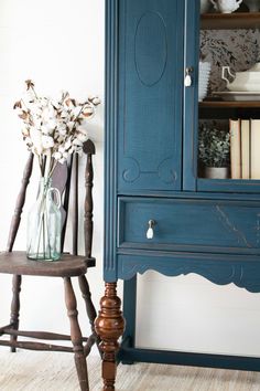 an old chair next to a blue china cabinet with flowers in the vase on top