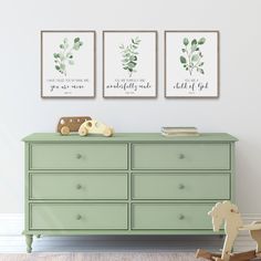 three framed art prints on the wall above a dresser with a rocking horse and baby's crib