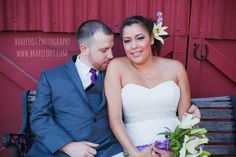 a bride and groom sitting on a bench in front of a red barn with flowers