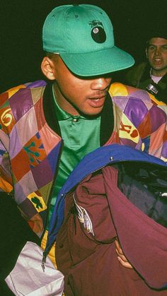 a man in a colorful jacket and hat is looking down at his backpack while another man stands behind him