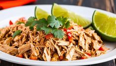 a white plate topped with shredded chicken and garnish next to lime wedges