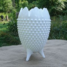 a large white vase sitting on top of a cloth covered tablecloth with trees in the background